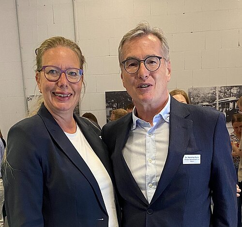 Eine Frau, Nicole Mölling (links), und ein Herr, Dr. Henning Rauls, stehen in Businesskleidung nebeneinander und lächeln in die Kamera. Im Hintergrund sind die Teilnehmer der Veranstaltung nur sehr vage zu erkennen.