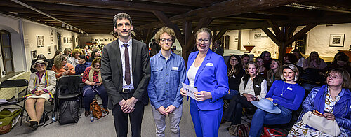 Drei Personen posieren vor einem Publikum in einem Raum mit Holzbalken. Im Hintergrund sitzt ein weibliches Publikum.
