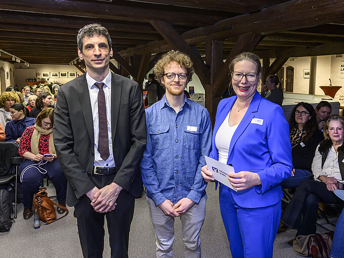 Drei Personen posieren vor einem Publikum in einem Raum mit Holzbalken. Im Hintergrund sitzt ein weibliches Publikum.