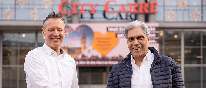 Zwei Männer schütteln sich die Hand vor einem Einkaufszentrum mit einem Schild von "City Carré" im Hintergrund.