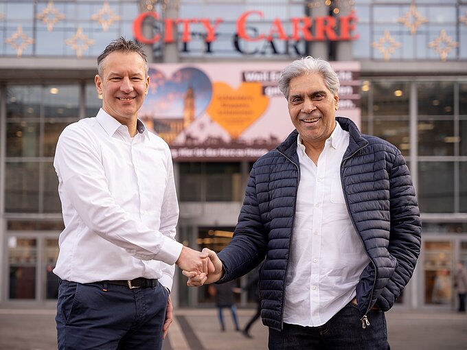 Zwei Männer schütteln sich die Hand vor einem Einkaufszentrum mit einem Schild von "City Carré" im Hintergrund.
