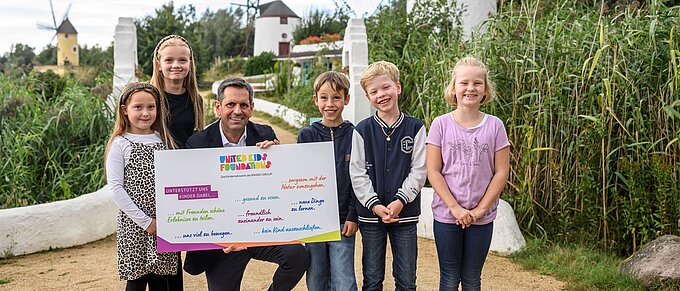 Eine Gruppe von fünf Kindern und einem Erwachsenen steht auf einem Weg in einem grünen, ländlichen Gebiet mit im Hintergrund sichtbaren Windmühlen. Der Erwachsene hält ein großes Schild mit bunten Texten, das das Logo der "United Kids Foundations" trägt. Alle Personen lächeln und wirken fröhlich. Die Umgebung ist von grünen Pflanzen und Sträuchern geprägt, und der Himmel ist teils bewölkt.