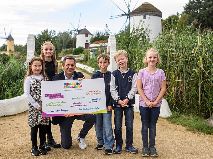 Eine Gruppe von fünf Kindern und einem Erwachsenen steht auf einem Weg in einem grünen, ländlichen Gebiet mit im Hintergrund sichtbaren Windmühlen. Der Erwachsene hält ein großes Schild mit bunten Texten, das das Logo der "United Kids Foundations" trägt. Alle Personen lächeln und wirken fröhlich. Die Umgebung ist von grünen Pflanzen und Sträuchern geprägt, und der Himmel ist teils bewölkt.
