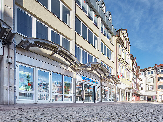 Straßenansicht mit Volksbank-Filiale und kik-Geschäft im Erdgeschoss eines mehrstöckigen Gebäudes bei blauem Himmel.