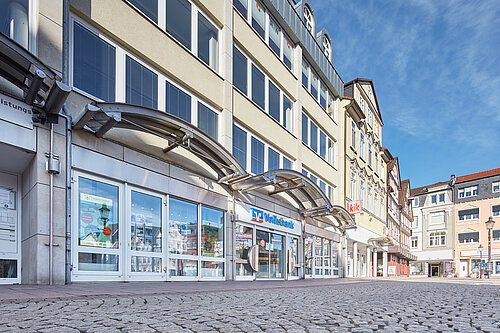 Straßenansicht mit Volksbank-Filiale und kik-Geschäft im Erdgeschoss eines mehrstöckigen Gebäudes bei blauem Himmel.