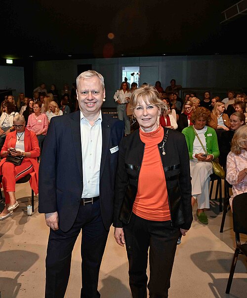 Auf dem Bild sieht man einen Mann und eine Frau im Anzug, die vor einem großen Publikum in einer Halle stehen. Der Mann trägt ein weißes Hemd und die Frau eine orangefarbene Bluse.
