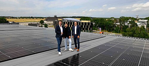 Drei Männer stehen auf einem Dach voller Solarpanels mit Gebäuden und Landschaft im Hintergrund.