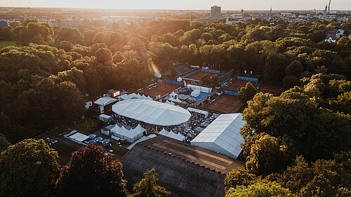 Luftaufnahme eines Tennisgeländes mit Zelten, umgeben von Bäumen und Gebäuden bei Sonnenuntergang.