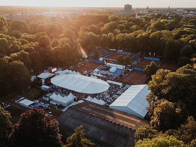 Luftaufnahme eines Tennisgeländes mit Zelten, umgeben von Bäumen und Gebäuden bei Sonnenuntergang.