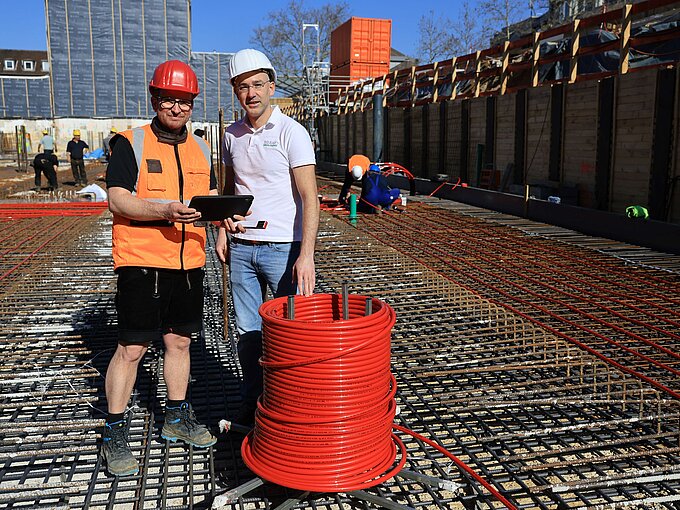 Bauarbeiter auf Baustelle mit Helm und Tablet vor Stahlkonstruktion, rote Kabelspule im Vordergrund.