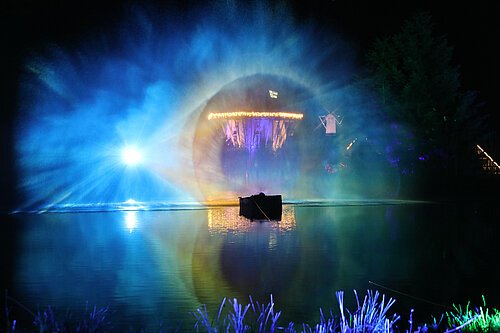 Buntes Lichtspektakel über einem nächtlichen See mit einem beleuchteten Gebäude im Hintergrund.