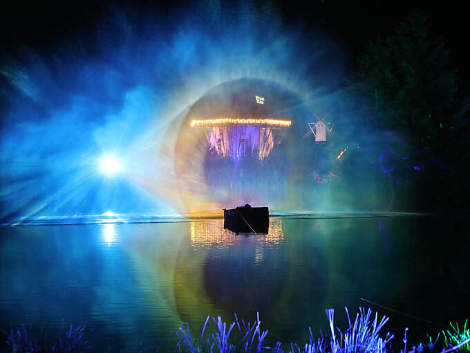 Buntes Lichtspektakel über einem nächtlichen See mit einem beleuchteten Gebäude im Hintergrund.