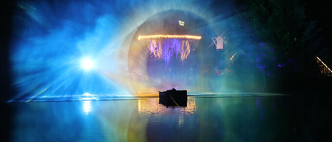 Buntes Lichtspektakel über einem nächtlichen See mit einem beleuchteten Gebäude im Hintergrund.