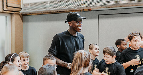 Dennis Schröder in einem schwarzen Shirt und einer Mütze lacht und interagiert fröhlich mit einer Gruppe von Kindern in Sportkleidung.