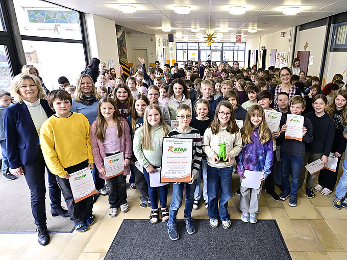 Große Gruppe von Kindern in einem Schulflur mit Zertifikaten und einem grünen Pokal, begleitet von Erwachsenen.