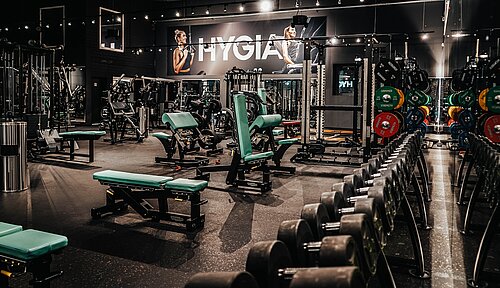 Leerer Fitnessraum mit modernen Geräten, Hanteln und Gewichten, grünen Sitzbänken und großen Wandpostern.