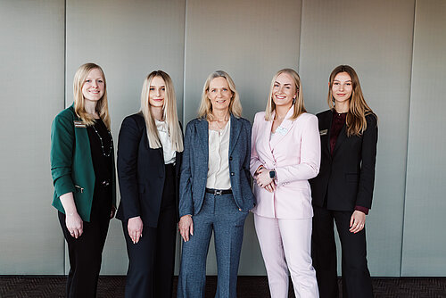 Auf dem Foto sieht man fünf Frauen in Anzügen, die nebeneinander stehen und in die Kamera lächeln. 