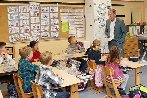 Ein Mann im Anzug steht in einem Klassenzimmer und spricht zu einer Gruppe Grundschulkinder, die aufmerksam an ihren Tischen sitzen. An der Wand hängen bunte Lernplakate mit englischen Vokabeln und Klassenzimmerausdrücken. Die Atmosphäre wirkt konzentriert und freundlich.