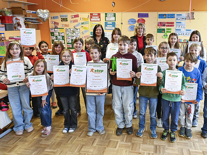 Kinder in einem Klassenzimmer halten stolz Zertifikate und posieren lächelnd, Erwachsene stehen dahinter.
