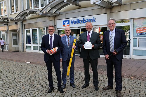 Vier Männer in Anzügen vor einer Volksbank, halten Bauutensilien wie Helm und Wasserwaage.