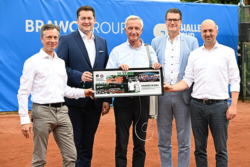 Auf dem Foto sieht man 5 Männer in Hemden und Anzügen, die vor einer blauen Wand auf einem Tennisplatz stehen und ein gerahmtes Bild hochhalten auf dem "30 Years" steht.