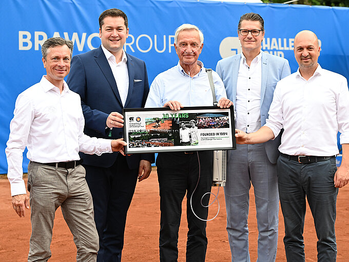 Auf dem Foto sieht man 5 Männer in Hemden und Anzügen, die vor einer blauen Wand auf einem Tennisplatz stehen und ein gerahmtes Bild hochhalten auf dem "30 Years" steht.