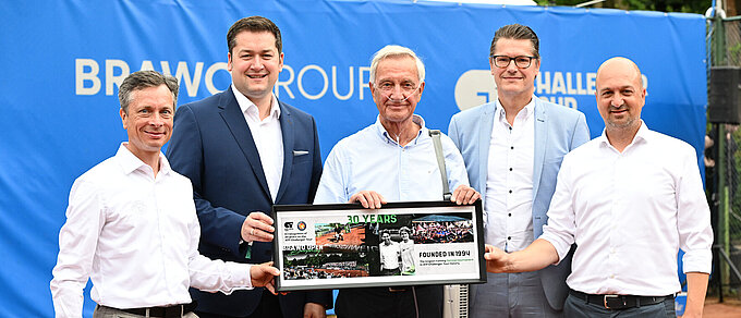 Auf dem Foto sieht man 5 Männer in Hemden und Anzügen, die vor einer blauen Wand auf einem Tennisplatz stehen und ein gerahmtes Bild hochhalten auf dem "30 Years" steht.