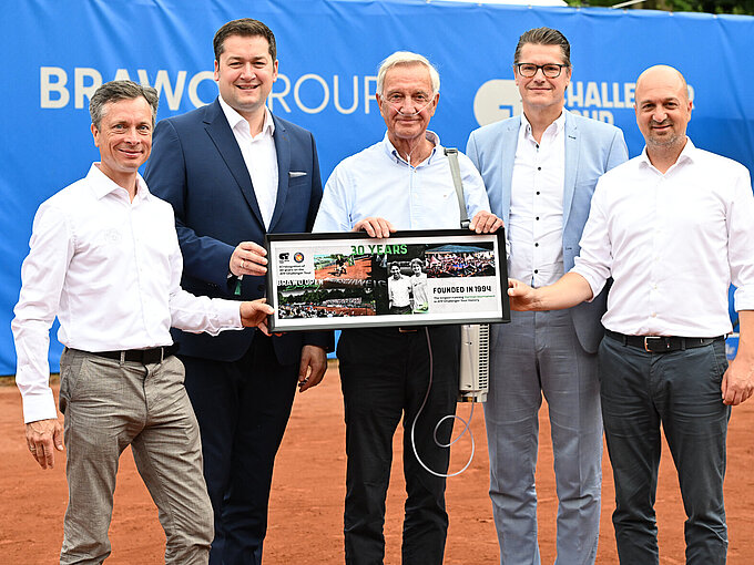 Auf dem Foto sieht man 5 Männer in Hemden und Anzügen, die vor einer blauen Wand auf einem Tennisplatz stehen und ein gerahmtes Bild hochhalten auf dem "30 Years" steht.