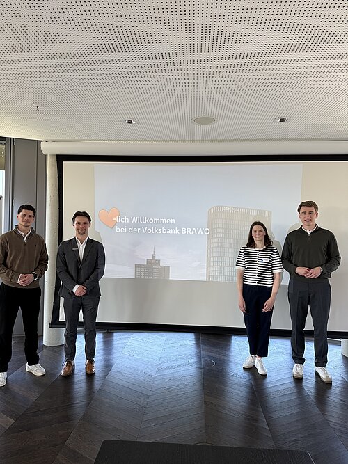 Vier Personen stehen vor einer Leinwand mit der Aufschrift "Willkommen bei der Volksbank BRAWO".