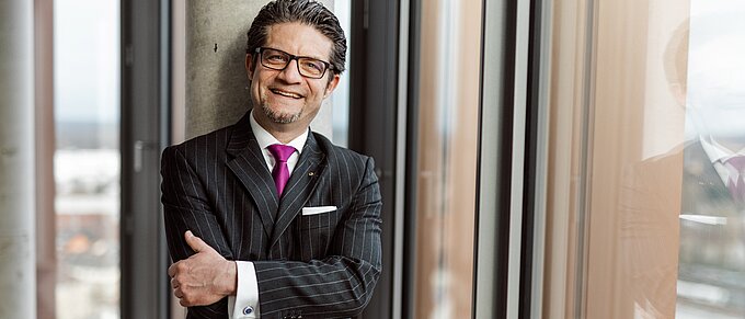 Mann im Anzug mit Krawatte steht lächelnd an einem Fenster, Arme verschränkt.