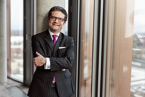 Mann im Anzug mit Krawatte steht lächelnd an einem Fenster, Arme verschränkt.