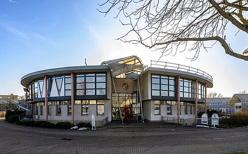 Modernes rundes Gebäude mit Glasfronten und Metallstruktur, umgeben von kahlen Bäumen an einem klaren Tag.