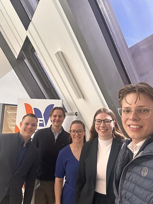 Fünf Personen lächeln in die Kamera vor einem modernen Fenster und einem Banner in einem hellen Raum.