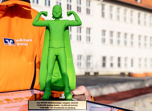 Grüne Superheldenfigur (Pokal) mit erhobenen Armen, gehalten von Person in orangener Jacke mit Logo der Volksbank BRAWO.