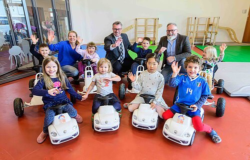 Kinder und Erwachsene posieren lächelnd und winkend in einer Kita mit Spielzeugautos.
