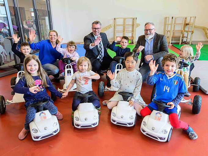 Kinder und Erwachsene posieren lächelnd und winkend in einer Kita mit Spielzeugautos.