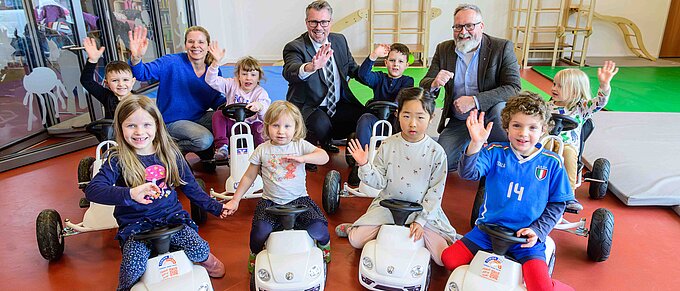 Kinder und Erwachsene posieren lächelnd und winkend in einer Kita mit Spielzeugautos.