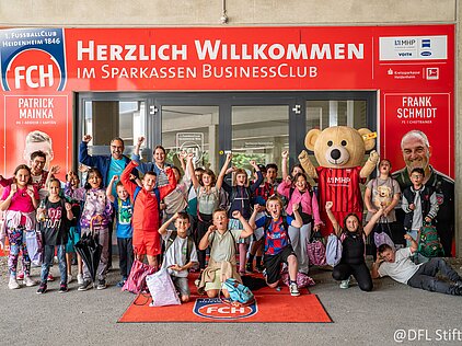 Kindergruppe jubelt mit Maskottchen vor dem Sparkassen BusinessClub des FC Heidenheim.