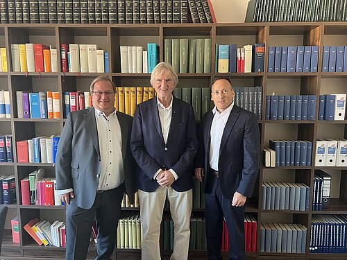 Drei Männer stehen nebeneinander in einem Raum vor einem großen Bücherregal, das voll mit vielen verschiedenen farbigen Gesetzesbüchern und Ordnern ist. Sie tragen Anzüge und lächeln in die Kamera. Die Atmosphäre wirkt professionell.