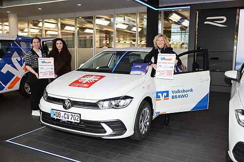 Drei Personen präsentieren ein VRmobil der Volksbank Brawo und Caritas mit Plakaten in der Autostadt in Wolfsburg.
