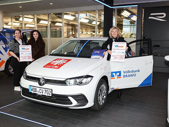 Drei Personen präsentieren ein VRmobil der Volksbank Brawo und Caritas mit Plakaten in der Autostadt in Wolfsburg.