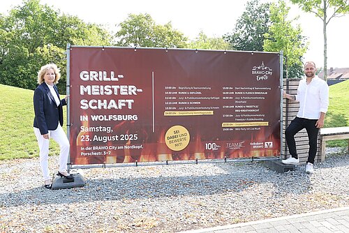 Zwei Personen stehen neben einem Bauzaunbannerplakat, die Frau links, der Mann rechts.