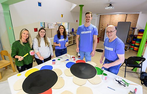 Eine Gruppe von fünf Personen steht an einem Tisch in einem Innenraum und malt große, blumenförmige Holzteile in verschiedenen Farben an. Einige tragen blaue T-Shirts, während andere in Alltagskleidung gekleidet sind. Auf dem Tisch liegen Farbgläser und Pinsel. Die Stimmung wirkt freundlich und gemeinschaftlich. Im Hintergrund sind bunte und hölzerne Elemente zu sehen.
