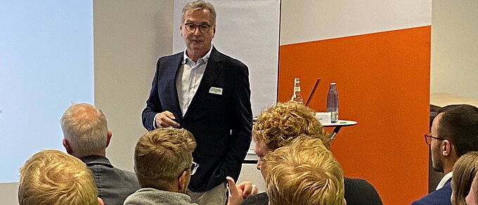 Im Hintergrund ist der Referent während seines Vortrags zu erkennen. Er schaut in die Runde der Teilnehmer der Veranstaltung, die auf dem Foto mit ihren Rücken und Hinterköpfen abgebildet sind.