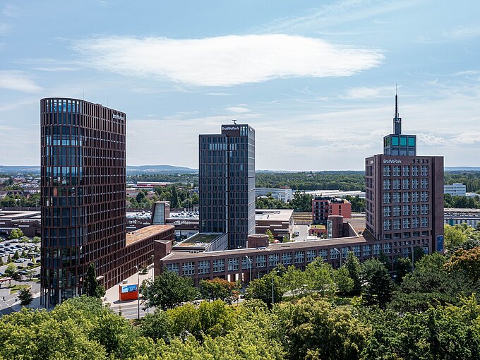 Stadtansicht mit modernen Hochhäusern, grünen Bäumen und einem klaren Himmel im Hintergrund.