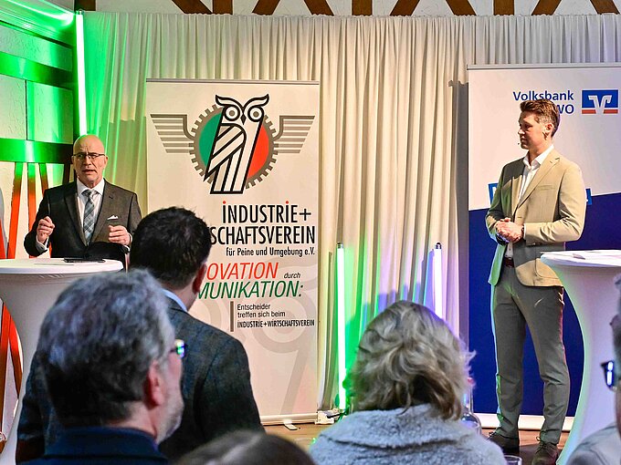 Zwei Männer halten Vorträge auf einer Veranstaltung mit Bannern des Industrie- und Wirtschaftsvereins und einer Bank.