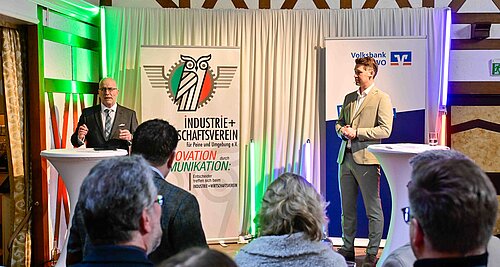 Zwei Männer halten Vorträge auf einer Veranstaltung mit Bannern des Industrie- und Wirtschaftsvereins und einer Bank.