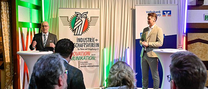 Zwei Männer halten Vorträge auf einer Veranstaltung mit Bannern des Industrie- und Wirtschaftsvereins und einer Bank.