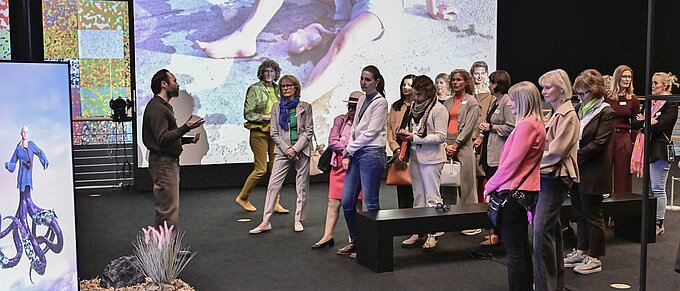 Person spricht vor einer Gruppe von Zuschauern in einer Galerie, Kunstwerke auf Bildschirmen im Hintergrund.