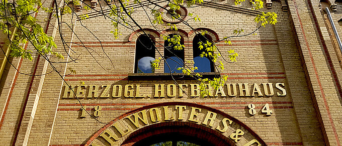 Backstein-Fassade des Herzog Hofbrauhauses mit goldener Schrift unter blauen Himmel und frischen grünen Blättern.